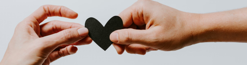 A close-up photograph capturing two pairs of hands, one on the left and one on the right, gently holding a single, solid black paper heart cut-out between their thumbs and forefingers against a plain, light grey background.