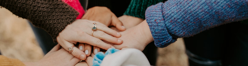 A close-up shot of several hands stacked on top of each other in a huddle, representing teamwork and unity. The individuals are wearing textured sweaters in shades of brown, teal, and blue, with one person wearing rings and white nail polish.